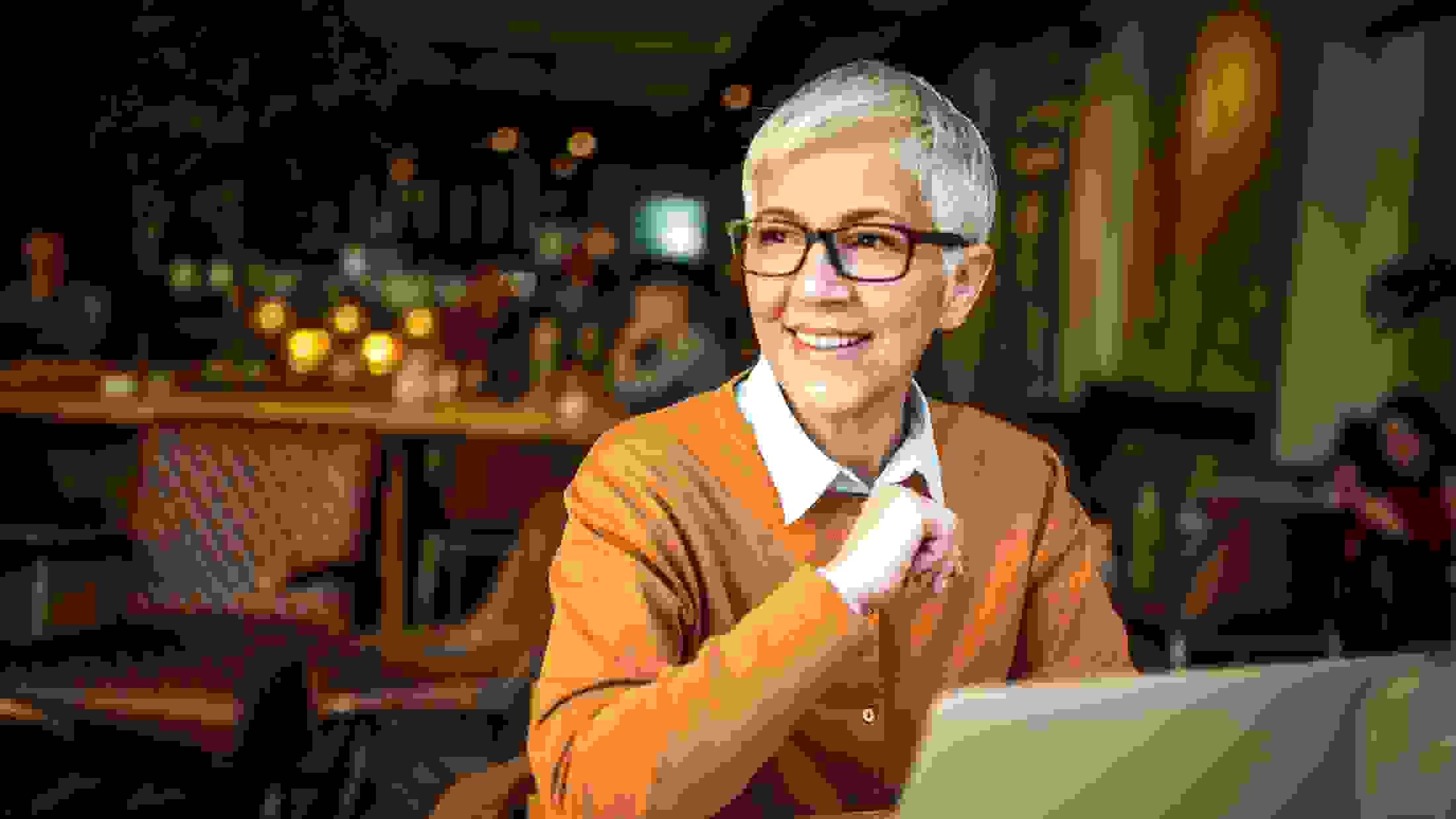 Middle aged woman with short hair and glasses wearing an orange jumper using her laptop while in a coffee shop