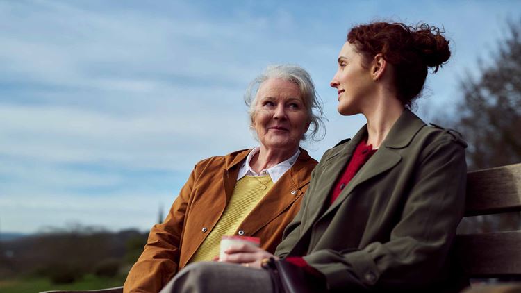 Mother and adult daughter sat on a bench drinking coffee