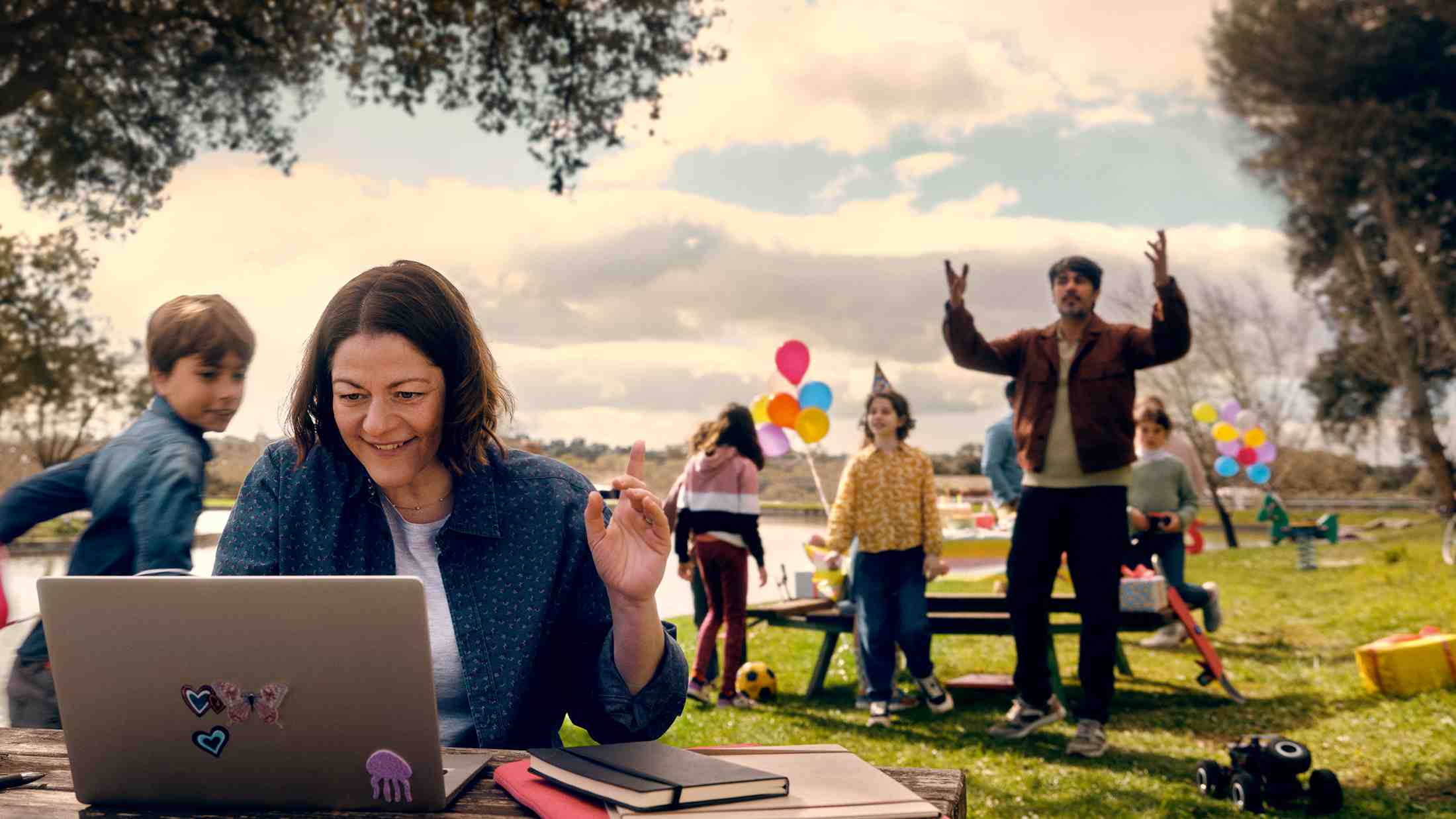 A female consultant working on her laptop outside on a nech while her family are having a birthday party in the background