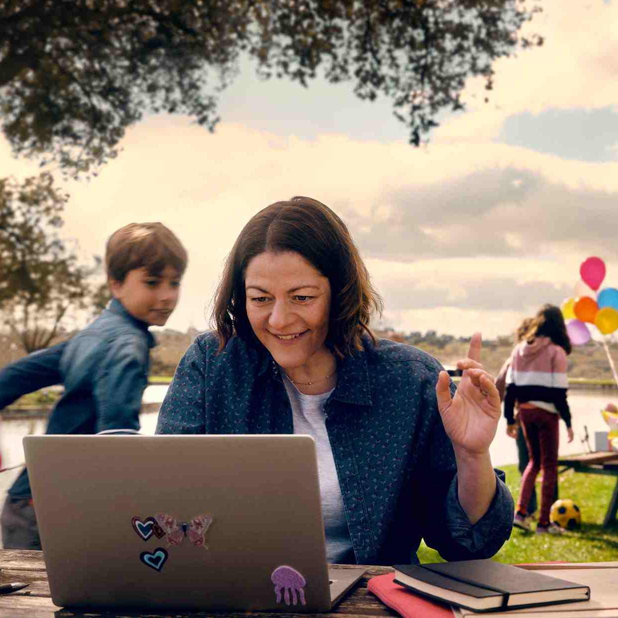 Female consultant working on her laptop outside on a bench while her family celebrate a birthday in the background