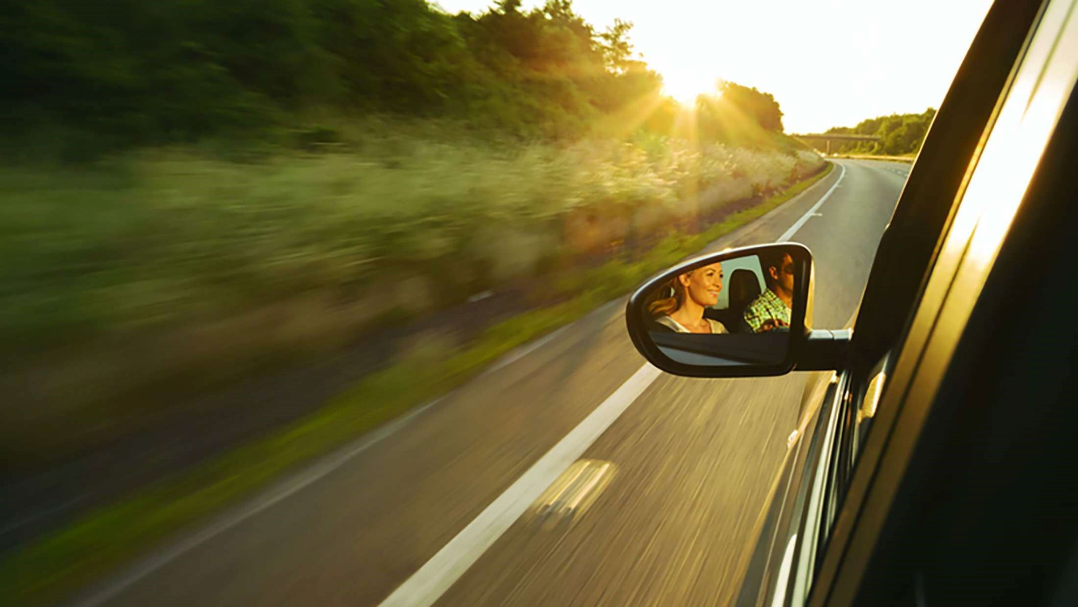 Car driving down road on a sunny evening with couple visible in the reflection of the left wing mirror