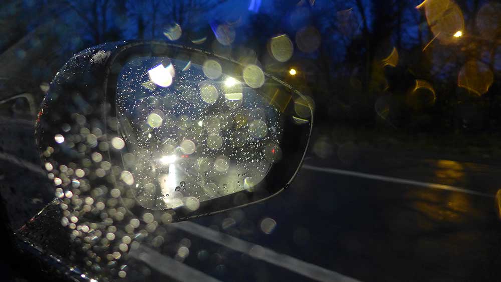 The side of a car at night with water droplets visible on the window and side mirror